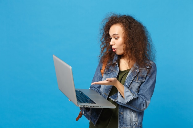 Young african american girl teen student in denim clothes, backpack work on pc isolated on blue wall