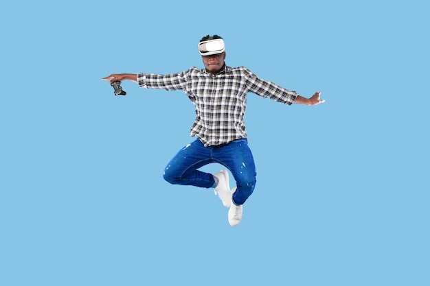 Young african american guy in vr headset holding joystick jumping up in air on blue background