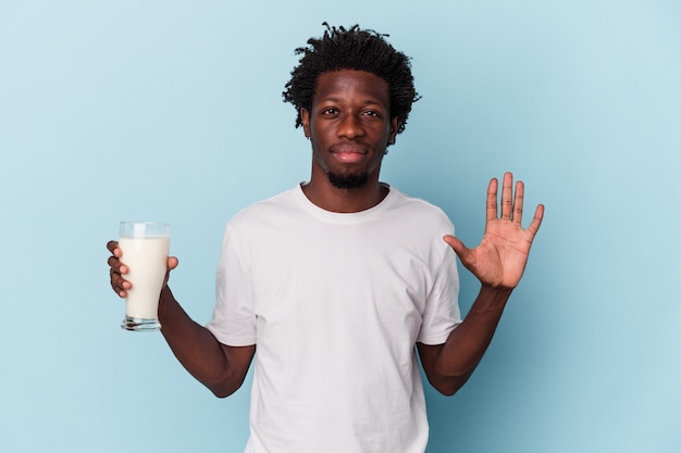 Young african american man holding a glass of milk isolated on blue background smiling cheerful showing number five with fingers.