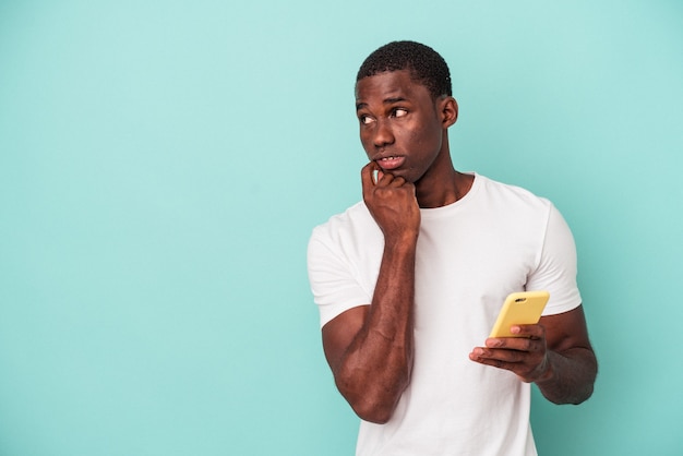 Young African American man holding a mobile phone isolated on blue background relaxed thinking about something looking at a copy space.