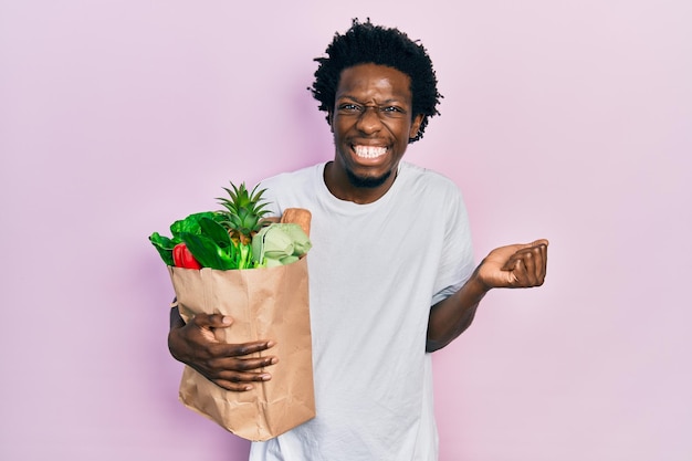 Young african american man holding paper bag with bread and groceries screaming proud, celebrating victory and success very excited with raised arm