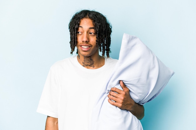 Young African American man holding a pillow isolated on blue background looks aside smiling cheerful and pleasant