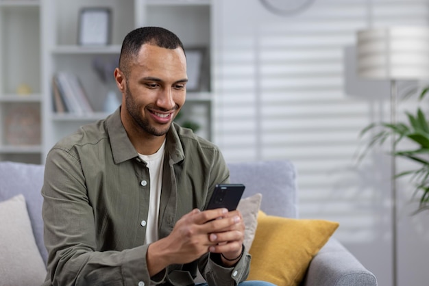 A young african american man is sitting on the couch at home and using the phone writes a message to