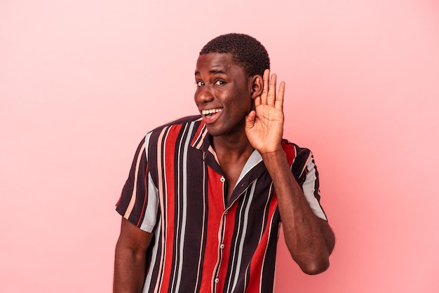 Young African American man isolated on pink background trying to listening a gossip