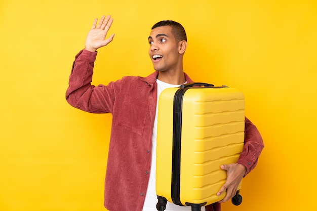 Young African American man over isolated yellow wall in vacation with travel suitcase and saluting