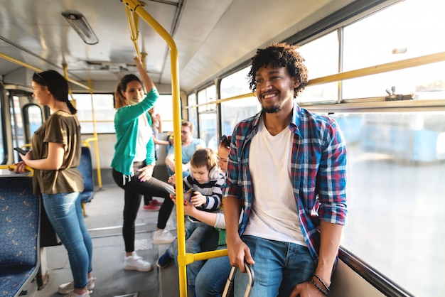 Young African American man standing in a bus and holding suitcase. In background people sitting and standing.