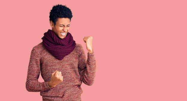 Young african american man wearing casual winter sweater and scarf celebrating surprised and amazed for success with arms raised and eyes closed winner concept