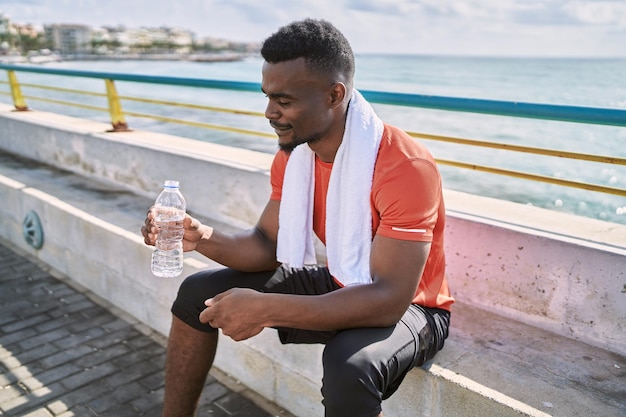 Young african american man wearing sportswear holding water bottle at seaside