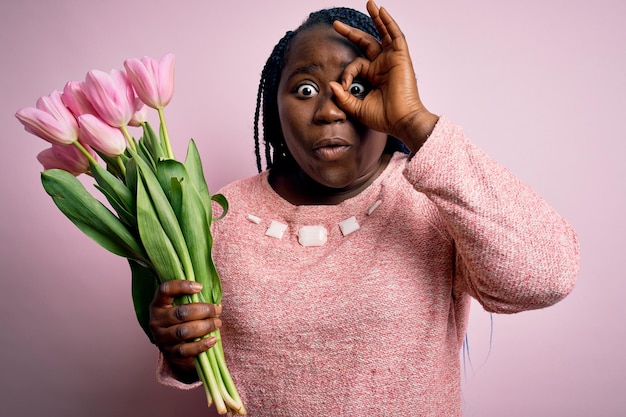 Young african american plus size woman with braids holding bouquet of pink tulips flower doing ok gesture shocked with surprised face eye looking through fingers Unbelieving expression