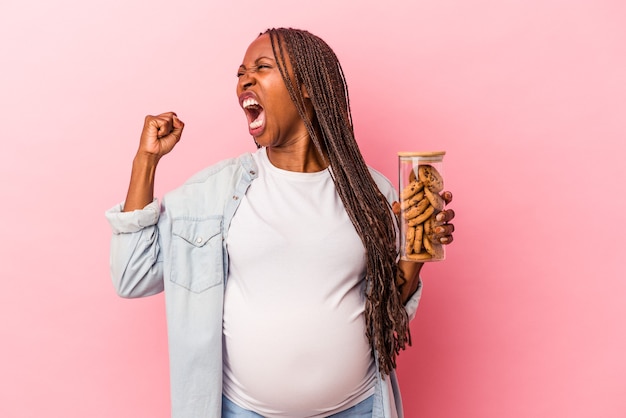 Young african american pregnant woman holding cookies isolated on pink background raising fist after a victory, winner concept.