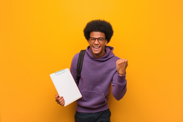 Young african american student man holding a clipboard surprised and shocked