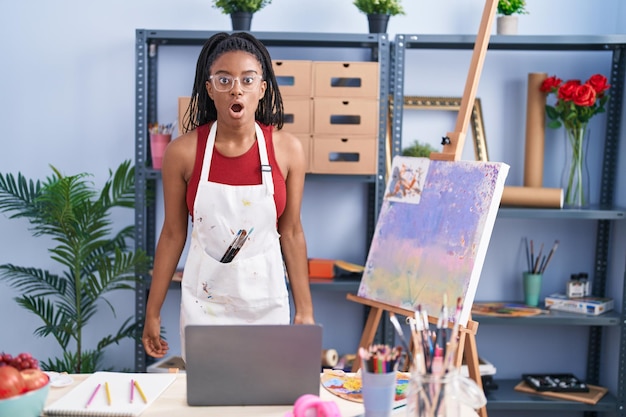 Young african american with braids at art studio painting on canvas scared and amazed with open mouth for surprise, disbelief face