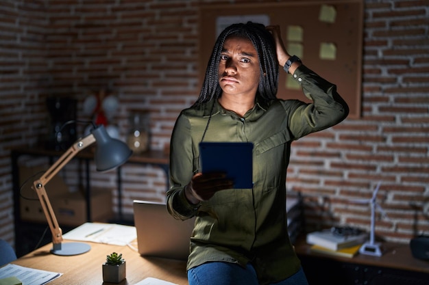 Young african american with braids working at the office at night confuse and wondering about question. uncertain with doubt, thinking with hand on head. pensive concept.