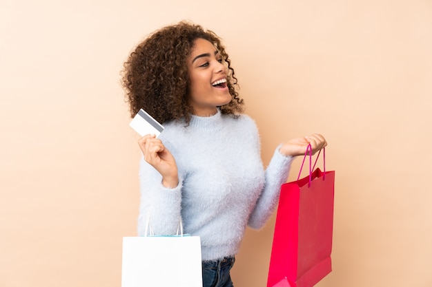 Young African American woman on beige wall holding shopping bags and a credit card