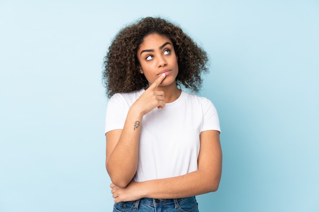 Young African American woman on blue wall having doubts while looking up