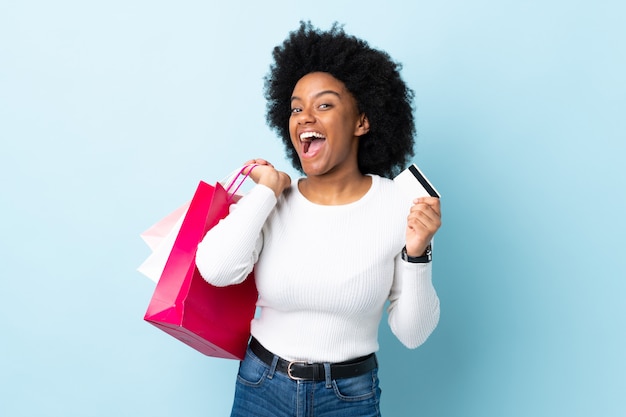 Young African American woman on blue wall holding shopping bags and a credit card