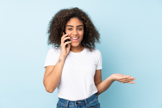 Young African American woman on blue wall keeping a conversation with the mobile phone with someone