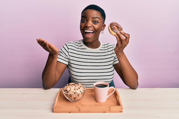 Young african american woman eating breakfast holding chocolate donut celebrating achievement with happy smile and winner expression with raised hand