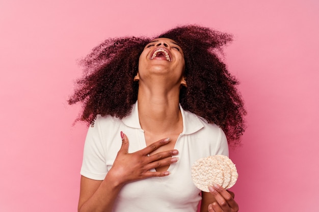 Young african american woman eating a rice cakes isolated on pink background laughs out loudly keeping hand on chest.