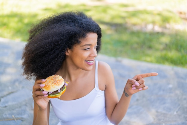 Young African American woman holding a burger at outdoors pointing to the side to present a product