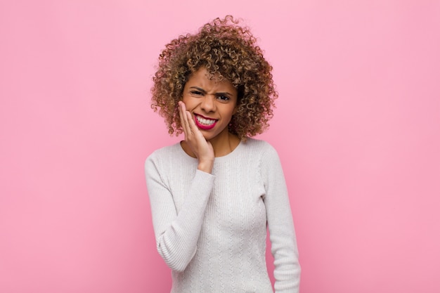 Young african american woman holding cheek and suffering painful toothache, feeling ill, miserable and unhappy, looking for a dentist against pink wall