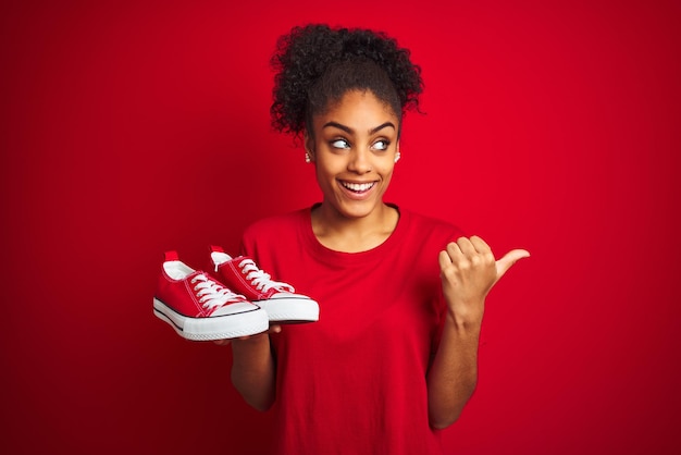 Young african american woman holding fashion sneakers over isolated red background pointing and showing with thumb up to the side with happy face smiling