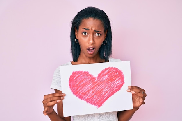 Young african american woman holding heart draw in shock face looking skeptical and sarcastic surprised with open mouth