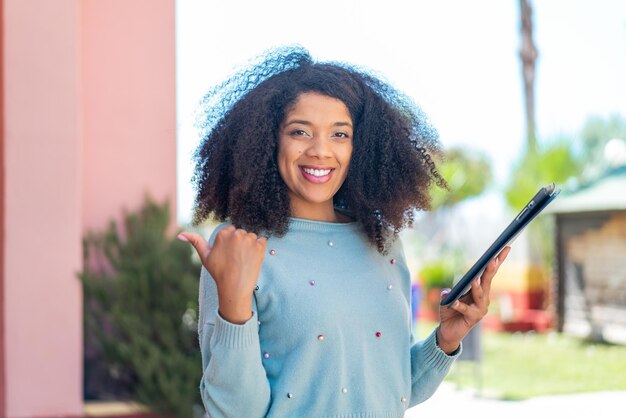 Young African American woman holding a tablet at outdoors pointing to the side to present a product