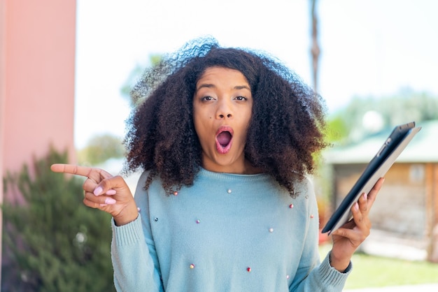 Young African American woman holding a tablet at outdoors surprised and pointing side