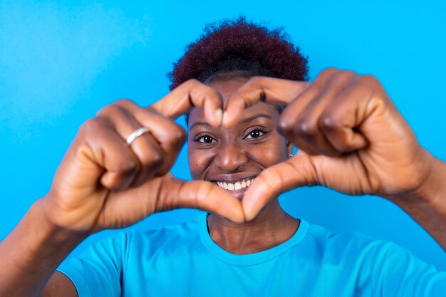 Young african american woman isolated on a blue background smiling and heart gesture studio shoot