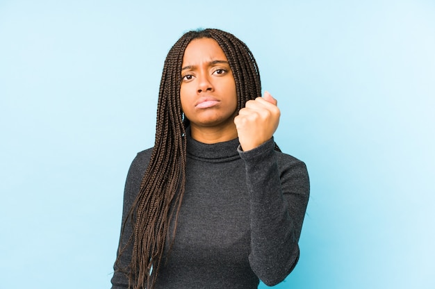 Young african american woman isolated on blue wall showing fist, aggressive facial expression.