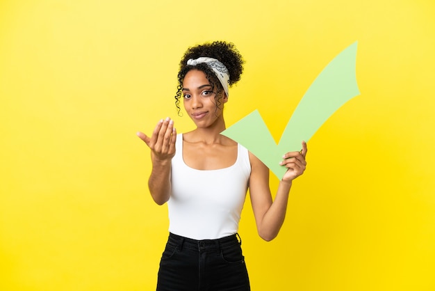 Young african american woman isolated on yellow background holding a check icon and doing coming gesture