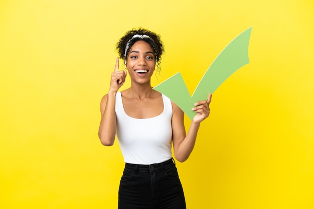 Young african american woman isolated on yellow background holding a check icon and pointing up