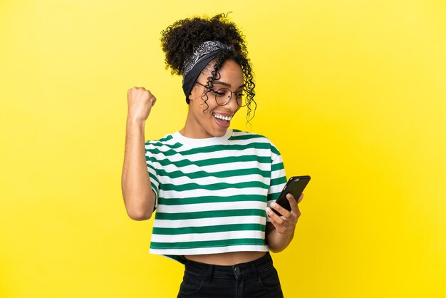 Young african american woman isolated on yellow background using mobile phone and doing victory gesture