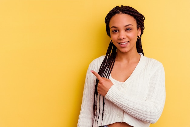 Young african american woman isolated on yellow wall smiling and pointing aside, showing something at blank space.