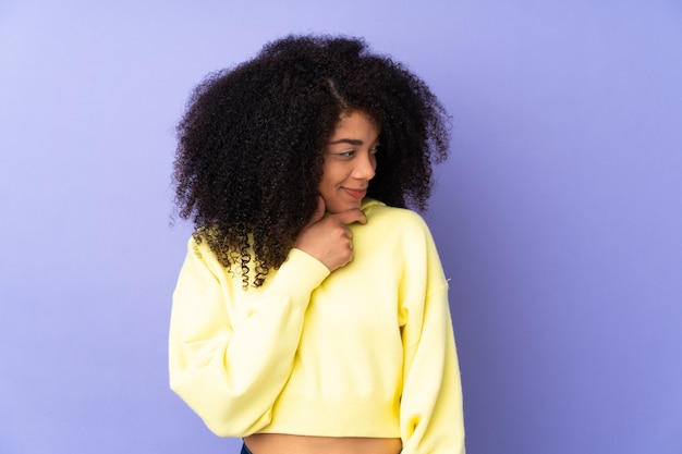 Young african american woman on purple wall looking to the side and smiling