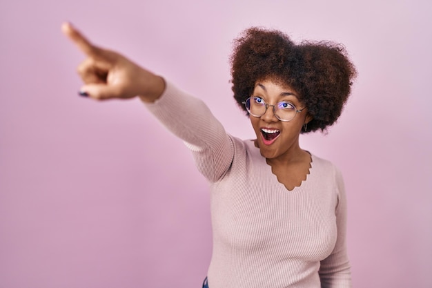 Young african american woman standing over pink background pointing with finger surprised ahead, open mouth amazed expression, something on the front