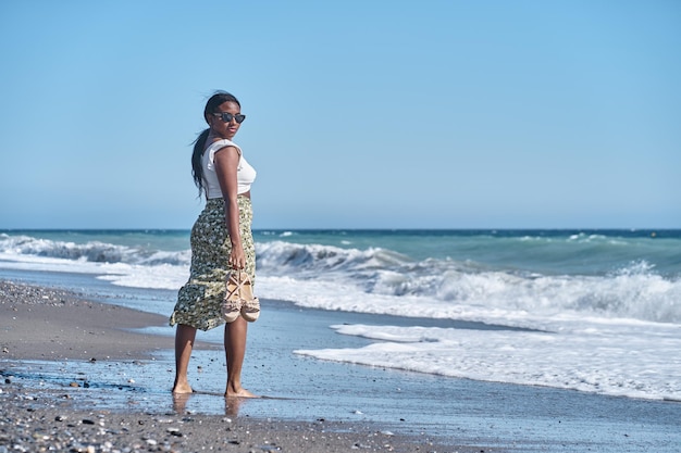 Young African American woman walking along the shore of the beach with sandals in hand