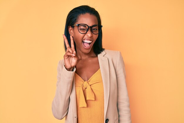 Young african american woman wearing business clothes smiling with happy face winking at the camera doing victory sign number two