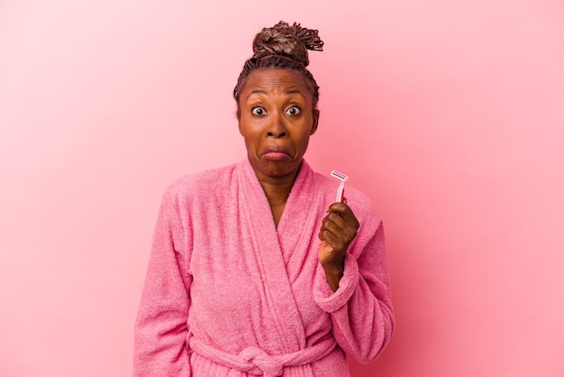 Young african american woman wearing pink bathrobe holding razor blade isolated on pink background shrugs shoulders and open eyes confused.