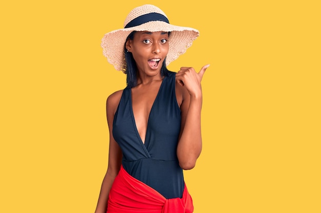 Young african american woman wearing swimsuit and summer hat smiling with happy face looking and pointing to the side with thumb up.