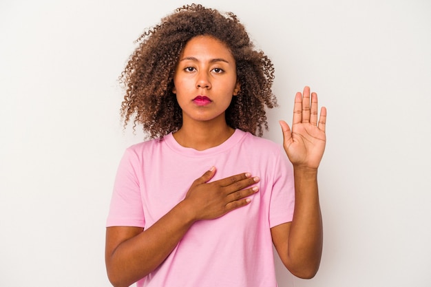 Young african american woman with curly hair isolated on white background taking an oath, putting hand on chest.