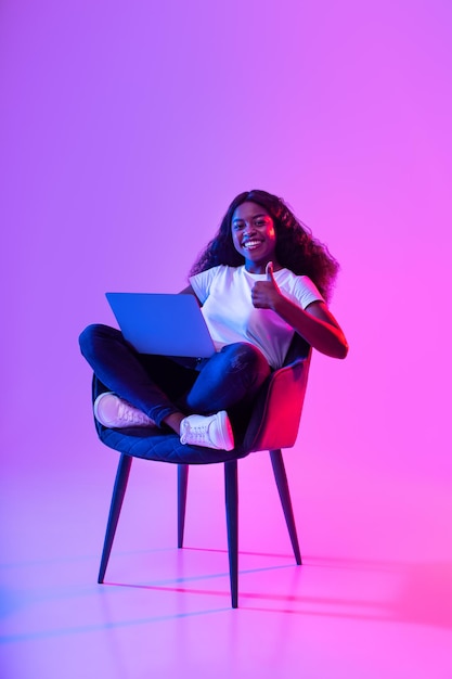 Young african american woman with laptop working online sitting in armchair and showing thumb up in