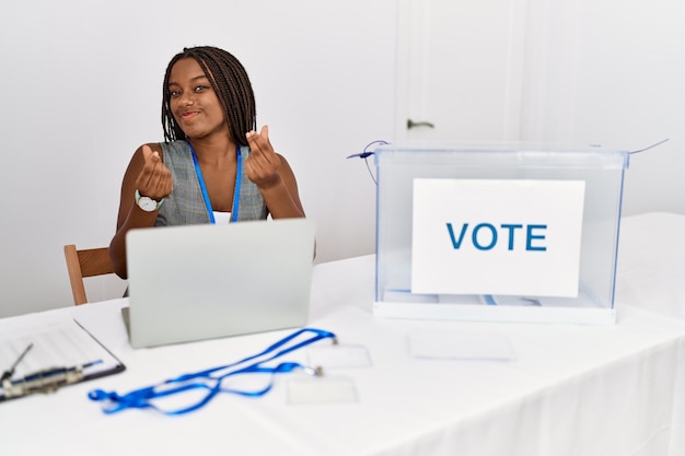 Young african american woman working at political election sitting by ballot doing money gesture with hands, asking for salary payment, millionaire business