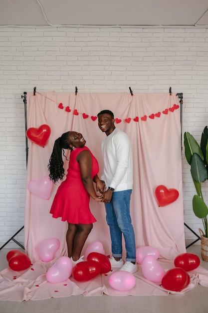 Young African couple holding hands while standing on pink background with heart shaped balloons