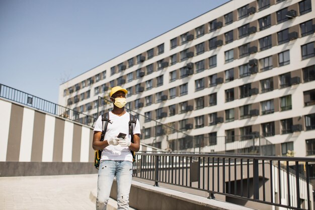 Young African courier in face mask walks down city street with thermal backpack