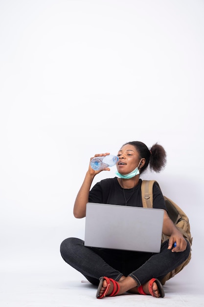 Young african lady carrying a backpack, sitting legs crossed using a laptop, drinking water