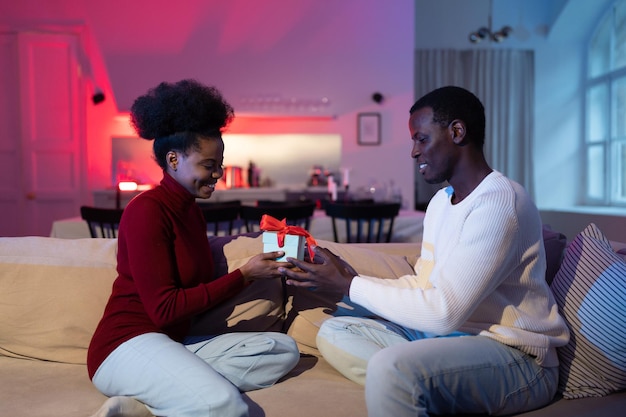 Young african man congratulates beloved woman with anniversary at home giving birthday gift box