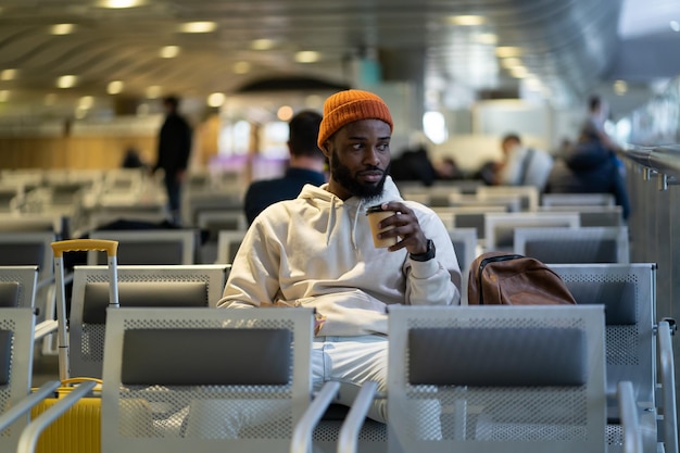 Young african man drinking coffee and eating sandwich while waiting for flying at airport terminal