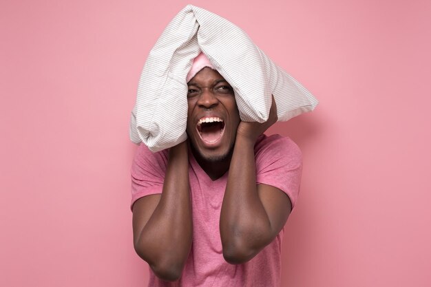 Young african man with pillow on head feeling good and amazed on pink wall